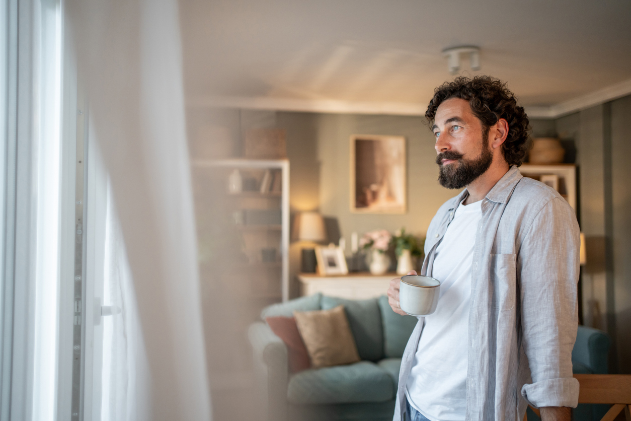 Mann i 40-årene står ettertenksom ved vinduet med en kaffekopp.
